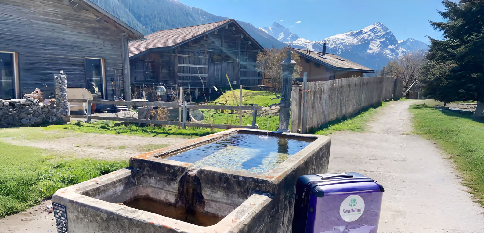 A rustic stone water trough filled with clear water sits outdoors next to a dirt path. In the foreground, a purple suitcase with a "Deafblind" logo is visible. Old wooden chalets and snow-capped mountains are in the background.