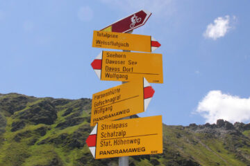 A yellow hiking signpost with multiple arrows pointing toward different trails (including Totalpsee, Weissfluhjoch, Davos Dorf, and Schatzalp) against a background of a steep, green mountain slope and a blue sky. A small maroon sign at the top shows a bicycle icon.