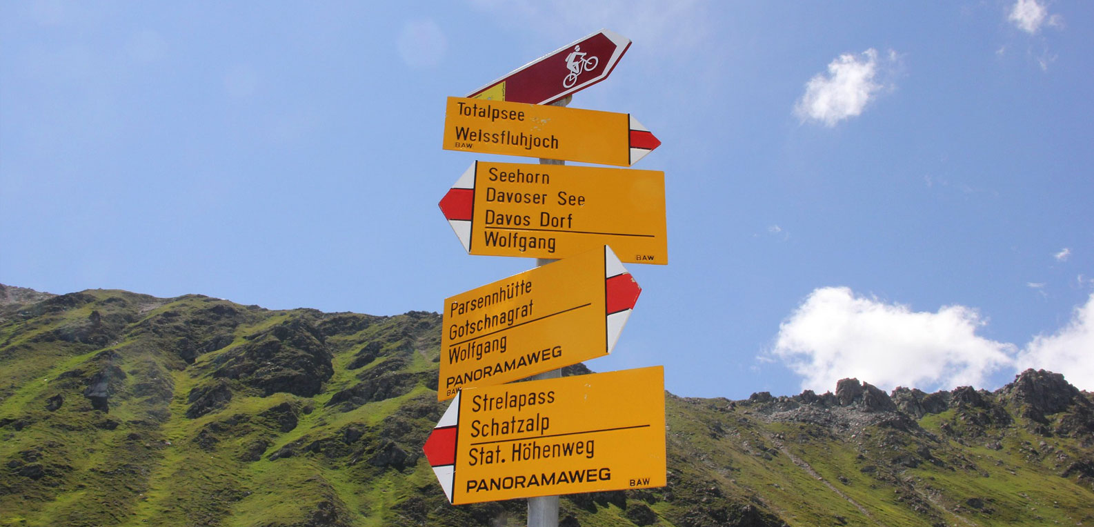 A yellow hiking signpost with multiple arrows pointing toward different trails (including Totalpsee, Weissfluhjoch, Davos Dorf, and Schatzalp) against a background of a steep, green mountain slope and a blue sky. A small maroon sign at the top shows a bicycle icon.